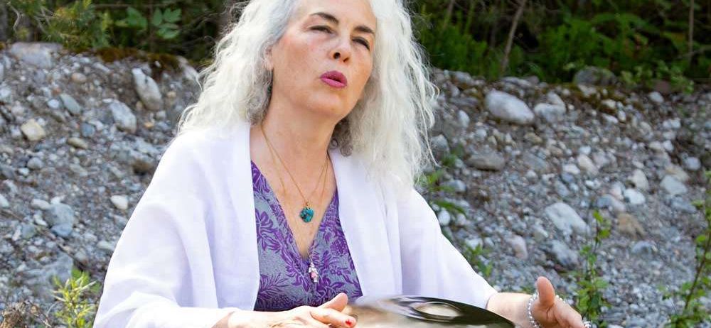 Eine ältere Frau mit weißen Haaren ist im Freien und hält eine große Metall-Handpan. Sie trägt einen weißen Umhang und ein lila Oberteil mit einer Halskette. Der Hintergrund ist ein felsiger Bereich mit Pflanzen.