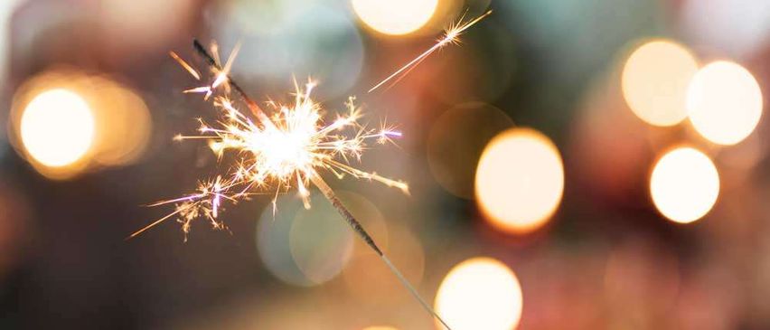 Eine Hand hält einen entzündeten Feuerwerkskörper, der einen funkelnden Hintergrund mit Bokeh-Lichtern beleuchtet. Der Feuerwerkskörper sendet helle, funkelnde Muster gegen einen festlichen, verblassten Hintergrund aus.