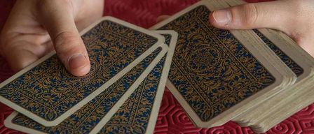Eine Person hält ein Deck mit verzierten Karten, die Finger sind über den Karten positioniert, vor einem strukturierten roten Hintergrund.