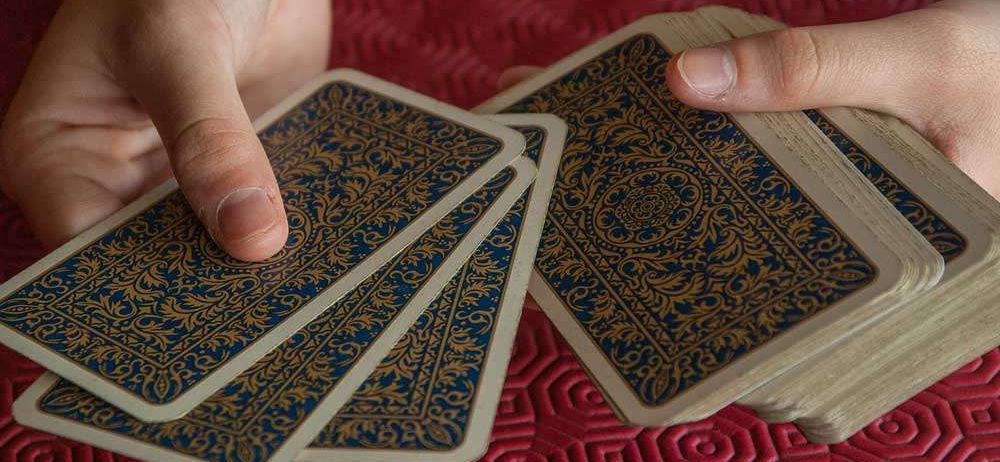 Eine Person hält ein Deck mit verzierten Karten, die Finger sind über den Karten positioniert, vor einem strukturierten roten Hintergrund.