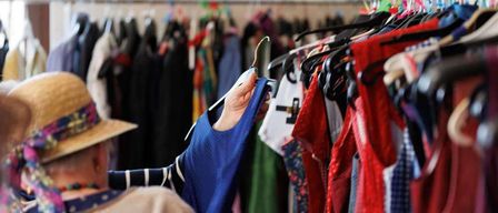 A person in a hat examines a blue shirt from a clothing rack. Various garments of different colors and styles hang on the rack.