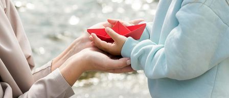 Zwei Hände, eine hält ein rotes Papierboot, sind vor einem ruhigen Gewässer eng beieinander.
