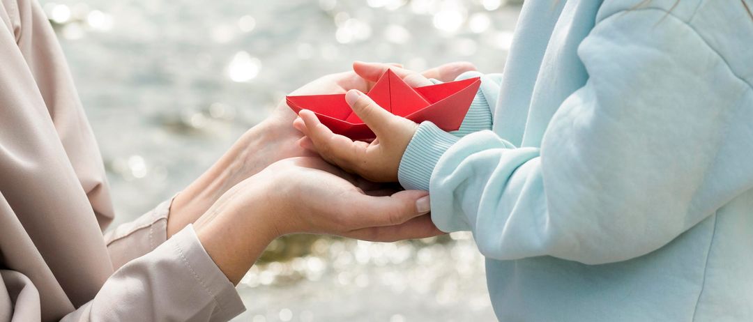 Zwei Hände, eine hält ein rotes Papierboot, sind vor einem ruhigen Gewässer eng beieinander.