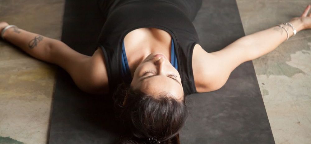 Eine Frau liegt auf einer schwarzen Yogamatte, mit ausgestreckten Armen und Beinen, die Augen geschlossen. Sie trägt ein schwarzes ärmelloses Top und scheint zu meditieren.