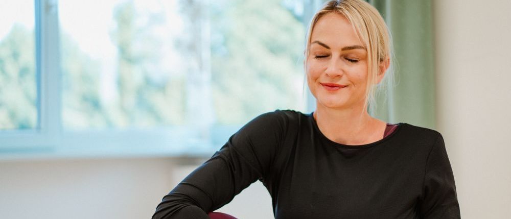Eine Frau mit blonden Haaren sitzt auf dem Boden, lächelt und scheint Yoga zu machen. Sie trägt ein schwarzes Langarmshirt und rote Hosen.
