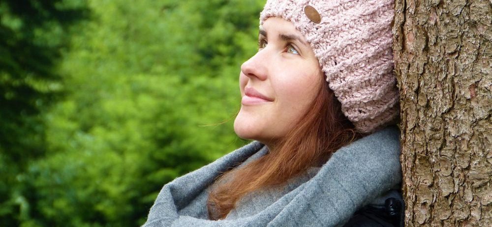 Eine Frau mit roten Haaren, die eine rosa Mütze und einen grauen Schal trägt, lehnt sich gegen einen Baum und lächelt in einem üppigen grünen Wald nach oben.