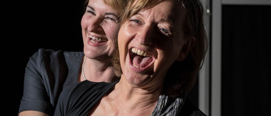 Zwei Frauen, eine mit einer Halskette, stehen eng beieinander, lachen und zeigen ihre Zähne, wobei eine Frau einen Schal um den Hals hat.