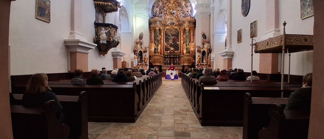 Eine Innenansicht einer Kirche mit Bänken, einem Altar und einer kunstvollen Wandgestaltung. Menschen sitzen auf den Bänken, und ein Priester steht am Altar.