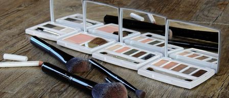 Eine Sammlung von Make-up-Pinseln und Lidschattenpaletten auf einer Holzoberfläche. Die Pinsel variieren in Größe und Form. Jede Lidschattenpalette enthält mehrere Farbtöne.