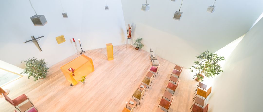 Ein leerer Raum mit Holzboden hat Reihen von Stühlen. Auf der linken Seite steht ein Tisch mit Kerzen. Rechts steht eine Topfpflanze.