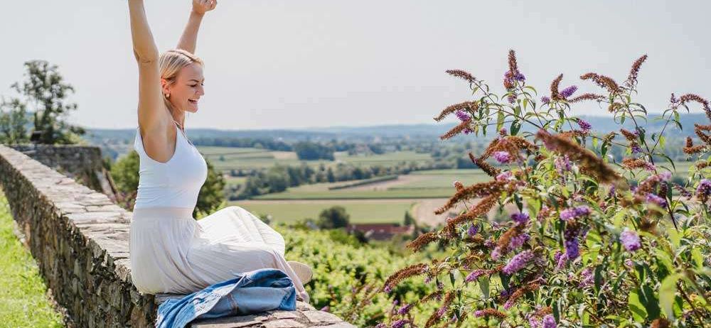 Eine Frau in einem weißen Kleid sitzt auf einer Steinmauer, hebt ihre Hände fröhlich in die Luft und lächelt. Sie wird von lila Blumen umgeben, und hinter ihr erstreckt sich ein weites Panorama einer grünen Landschaft.