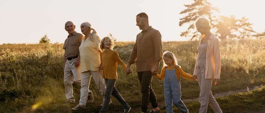 A family of seven walks through a grassy field as the sun sets. The adults and children hold hands.
