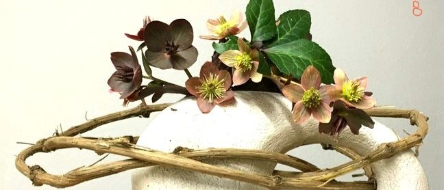 Eine weiße Keramikvase mit kreisförmiger Basis, geschmückt mit einem Zweig und verschiedenen Blumen. Die Vase steht auf einer Holzoberfläche.