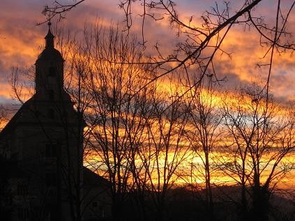 Eine Kirche mit einem Kirchturm ist vor einem Sonnenuntergang silhouettiert, mit kahlen Bäumen im Vordergrund.