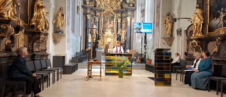 An altar in a church with two people standing in front. A table with flowers and a bowl is in the foreground. The room is adorned with statues and paintings.