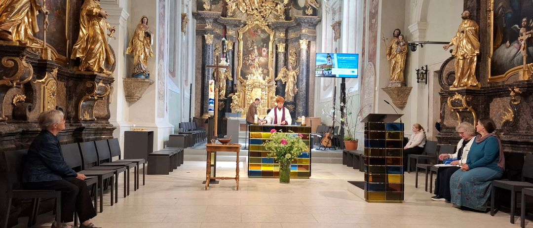 Ein Altar in einer Kirche mit zwei davor stehenden Personen. Ein Tisch mit Blumen und einer Schüssel im Vordergrund. Der Raum ist mit Statuen und Gemälden geschmückt.