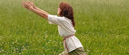A woman with a ring on her finger is standing in a grassy field and stretching her arms upwards.