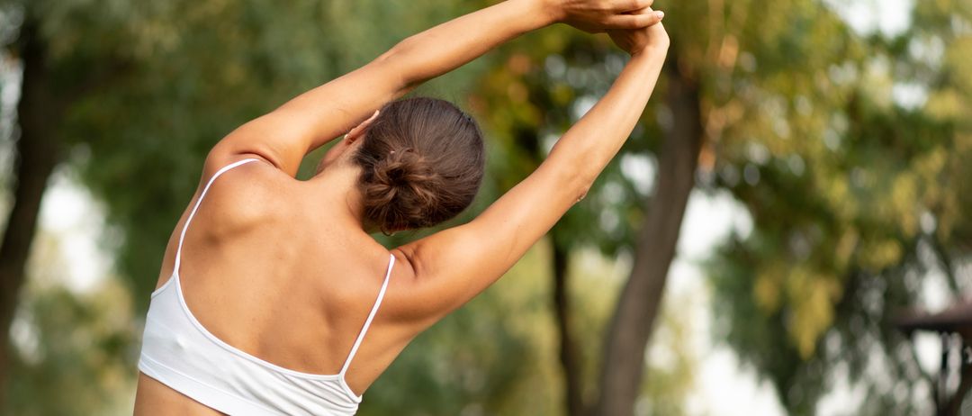 Eine Frau mit dem Rücken zur Seite, die ihre Arme in einem Park nach oben streckt. Sie trägt einen weißen Sport-BH und graue Hosen.