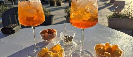 Zwei Gläser orangefarbener Getränke mit Strohhalmen, Schüsseln mit Pommes und Oliven auf einem weißen Tisch. Ein Auto und Gebäude sind im Hintergrund.