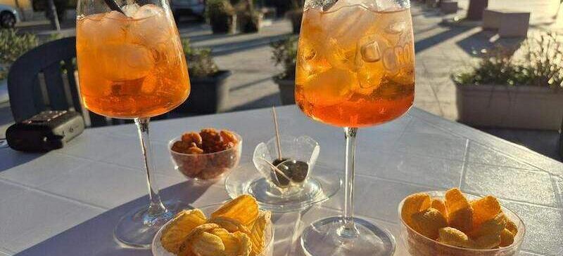 Two glasses of orange drinks with straws, bowls of chips, and olives on a white table. A car and buildings are in the background.