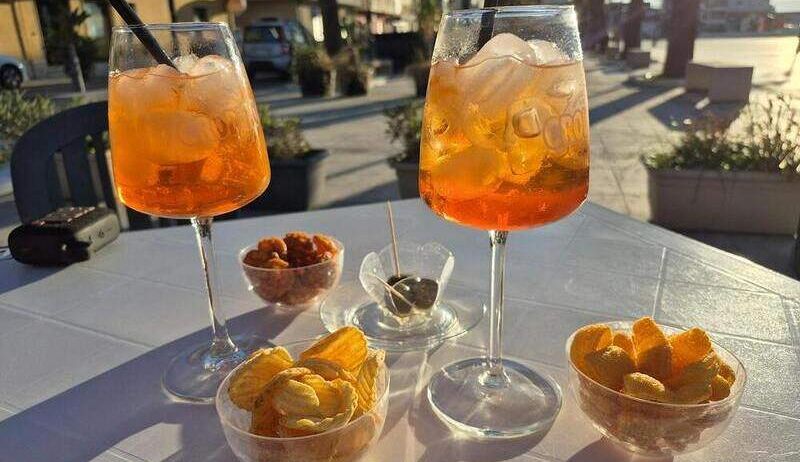 Zwei Gläser orangefarbener Getränke mit Strohhalmen, Schüsseln mit Pommes und Oliven auf einem weißen Tisch. Ein Auto und Gebäude sind im Hintergrund.