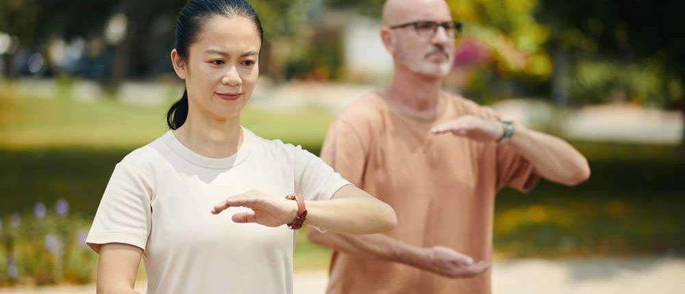 Eine Frau und ein Mann praktizieren Tai Chi im Freien, beide in Freizeitkleidung und lächelnd. Die Frau trägt ein weißes T-Shirt, der Mann ein pfirsichfarbenes Hemd.