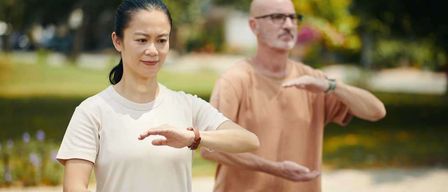 Eine Frau und ein Mann praktizieren Tai Chi im Freien, beide in Freizeitkleidung und lächelnd. Die Frau trägt ein weißes T-Shirt, der Mann ein pfirsichfarbenes Hemd.