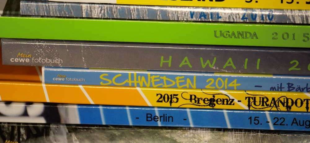 Ein Stapel von Reisefotobüchern mit Titeln wie 'Insel', 'Uganda', 'Hawaii', 'Schweden 2014', 'Bregenz-Tur' und 'Griechenland 2010'.