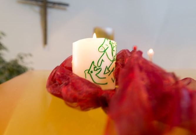 Eine Kerze mit einem grünen Design steht auf einem roten Tuch auf einem gelben Tisch. Ein Kreuz ist an der Wand hinter der Kerze angebracht.