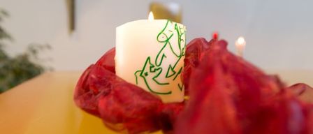 Eine Kerze mit einem grünen Design steht auf einem roten Tuch auf einem gelben Tisch. Ein Kreuz ist an der Wand hinter der Kerze angebracht.