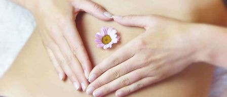 Die Hände einer Person bilden eine Herzform um eine rosa Blume auf ihrem Bauch.