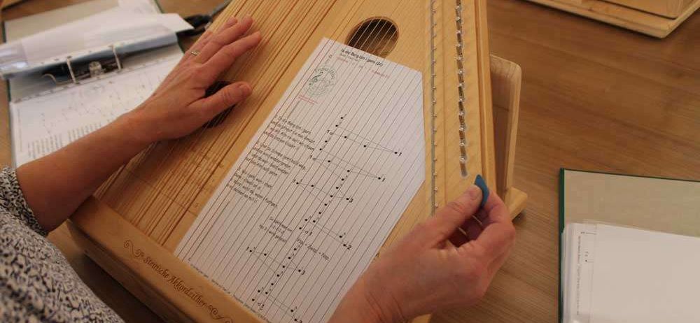 Eine Person spielt eine Zither mit einem Notenblatt darauf, sitzt an einem Holzschreibtisch.