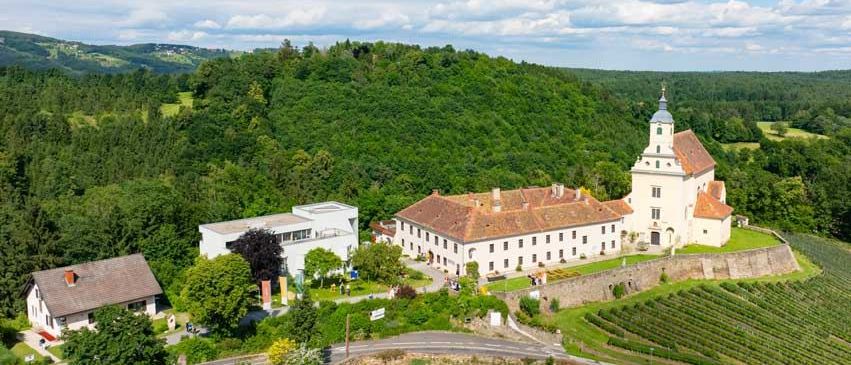 Eine Luftaufnahme eines historischen Gebäudes, möglicherweise eines Schlosses, auf einem Hügel umgeben von einem grünen Feld, Bäumen und Bergen.