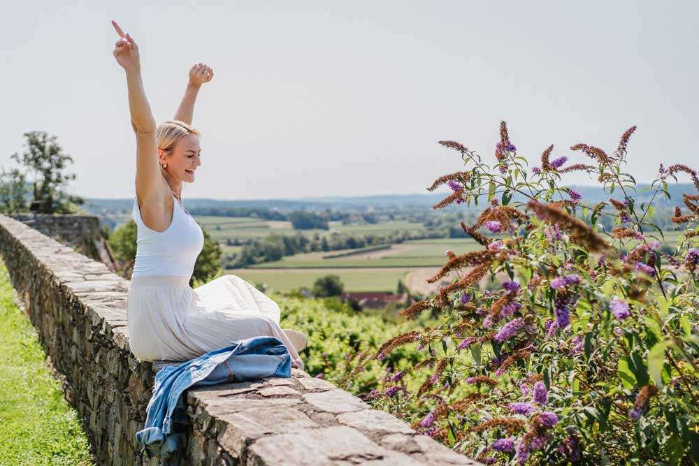 Eine Frau in einem weißen Kleid sitzt mit erhobenen Armen auf einer Steinmauer und blickt auf eine Landschaft mit Feldern und Bäumen.