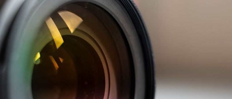 Nahaufnahme eines Kameraobjektivs mit sichtbaren Blendenlamellen und internen Elementen, die Licht in verschiedenen Farben einfangen.