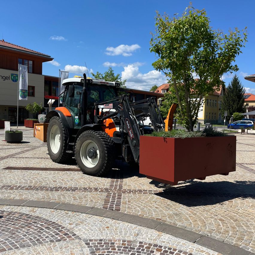 Ein orangefarbener Traktor zieht eine rote Blumenbank mit grünen Pflanzen und Gras auf einem Kopfsteinpflasterplatz vor einem Gebäude mit einer Flagge.