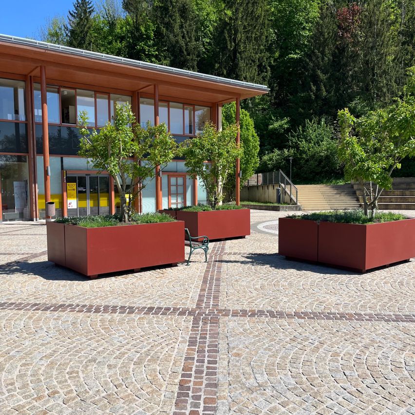 Außenbereich mit roten Pflanzgefäßen, einer grünen Bank und einem Gebäude mit Glasfenstern.