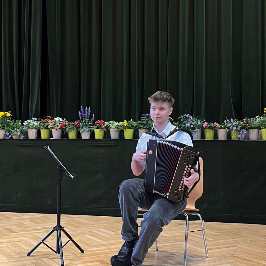 Ein junger Mann sitzt auf einem Stuhl und spielt ein Akkordeon in einem Raum mit schwarzem Hintergrund. Blumentöpfe säumen die Bühne hinter ihm.