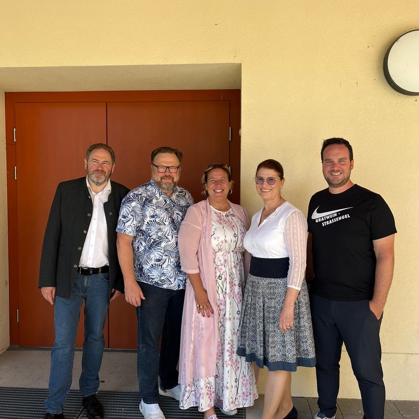 Fünf Personen, die lächeln und posieren, stehen vor einer roten Tür mit einer gelben Wand. Einer trägt ein Blumenhemd, ein anderer ein schwarzes Shirt mit Nike-Marke. Zwei Frauen tragen Kleider und eine trägt einen Blazer.