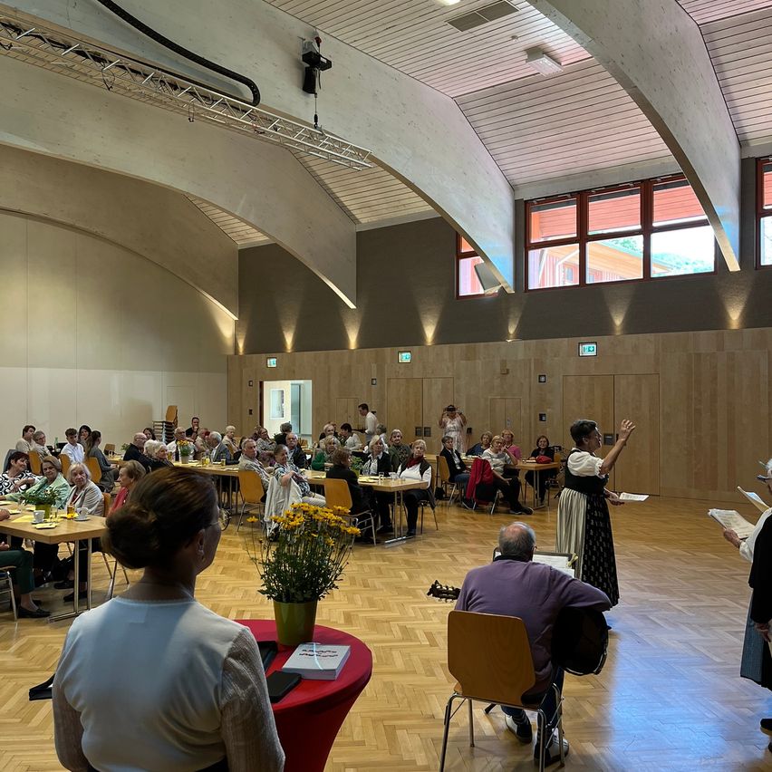 Eine Gruppe älterer Menschen versammelt sich in einem großen Raum. Sie sitzen an Tischen, und einige stehen. Vor ihnen steht eine Frau, die möglicherweise eine Versammlung leitet. Auf einem Tisch stehen Blumen, und ein Mann sitzt auf einem Stuhl mit einer Gitarre.