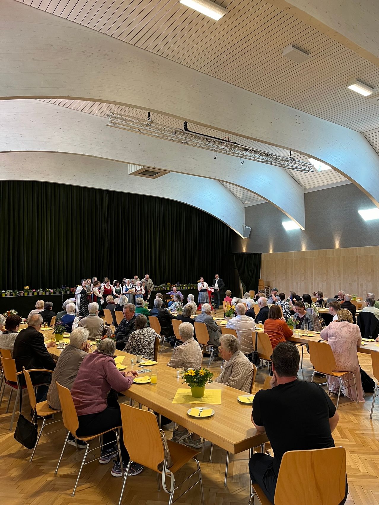 Eine Gruppe älterer Menschen sitzt an langen Tischen in einem Saal, wahrscheinlich bei einem gesellschaftlichen Zusammentreffen. Eine Bühne mit schwarzem Vorhang und einigen Personen ist vorne. Die Tische sind mit Blumen und Tellern geschmückt.