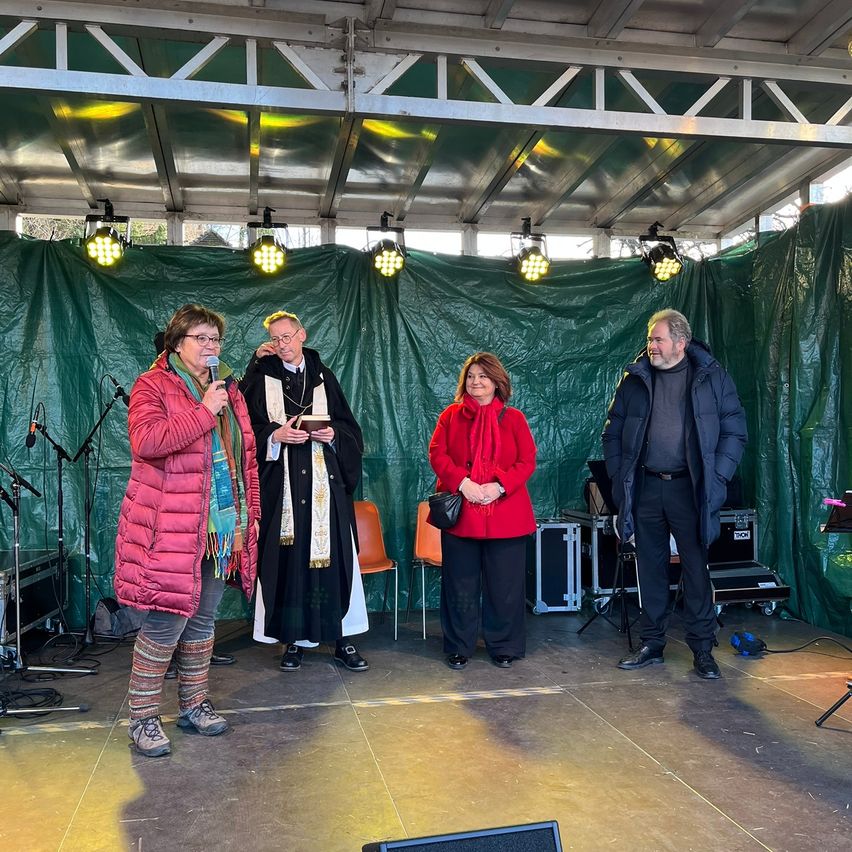 Vier Personen stehen auf einer Bühne mit grünem Hintergrund, Mikrofonen und Lautsprechern. Ein Mann in einem schwarzen Gewand hält ein Buch, während eine Frau ein Mikrofon hält. Eine andere Frau trägt einen roten Mantel, und ein Mann steht rechts.