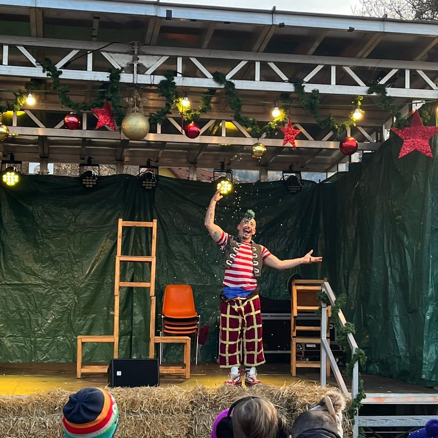Ein Clown tritt auf einer Bühne auf, die mit Weihnachtsdekorationen geschmückt ist, mit einem grünen Hintergrund und gestapelten Stühlen dahinter. Das Publikum schaut von unten zu.