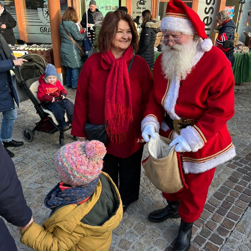 Der Weihnachtsmann unterhält sich mit einer Frau und einem Kind an einem festlichen Ort im Freien. In der Nähe ist ein Baby im Kinderwagen, und andere sind einkaufen.