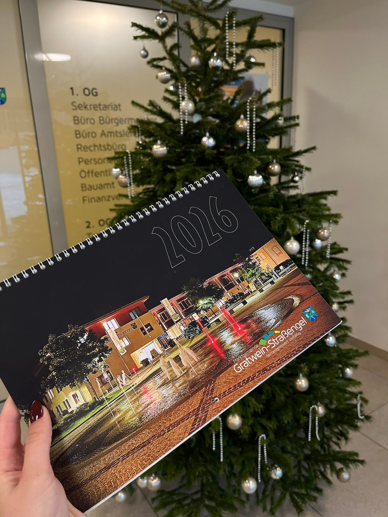 Eine Hand hält einen 2016 Kalender mit einer Stadtplatzansicht, auf der ein Brunnen im Vordergrund zu sehen ist. Dahinter ein Gebäude mit roten Mauern und Glasfenstern. Ein mit silbernen Kugeln geschmückter Weihnachtsbaum ist im Hintergrund.