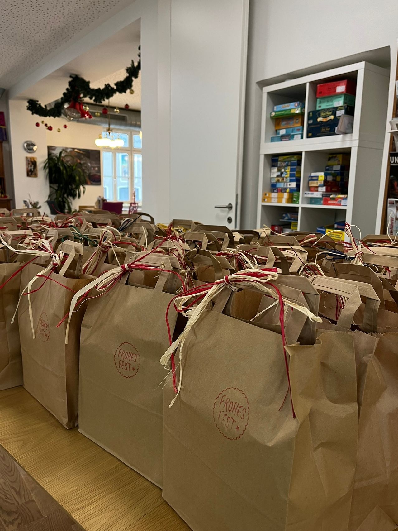 Ein Tisch mit vielen braunen Papiertüten mit Frohes Fest darauf, roten Bändern gebunden, und Regale mit verschiedenen Gegenständen im Hintergrund.