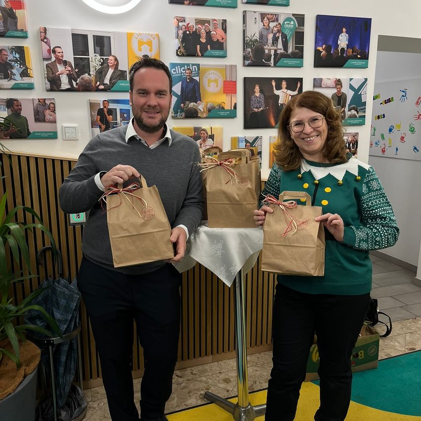 Ein Mann und eine Frau stehen in einem Raum mit einer Wand voller Bilder, lächeln und halten Papiertüten, die mit roten Bändern gebunden sind.