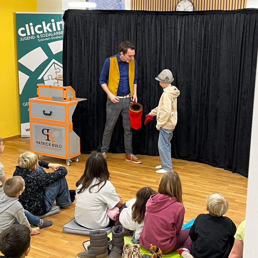 Ein Mann in einem blauen Hemd und einer gelben Weste steht auf der Bühne mit einem Jungen im Hut. Sie stehen vor einer schwarzen Kulisse mit einem Schild, das 'clickin' sagt. Mehrere Kinder sitzen vor ihnen auf dem Boden und schauen zu. Ein roter Eimer steht auf der Bühne.