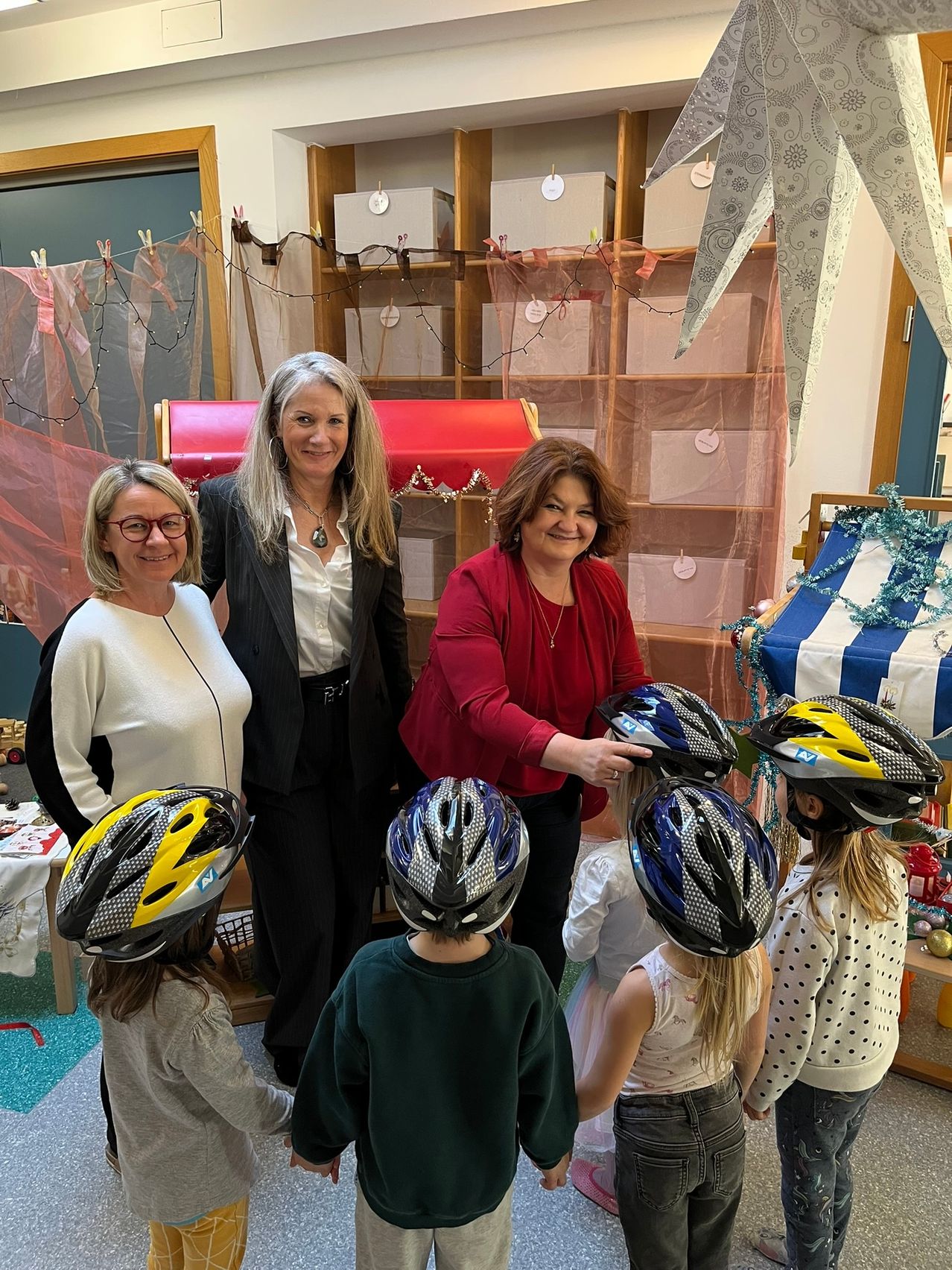 Mehrere Erwachsene und Kinder sind in einem Raum mit Regalen und Dekorationen. Eine Frau in einem roten Oberteil gibt den Kindern Helme.