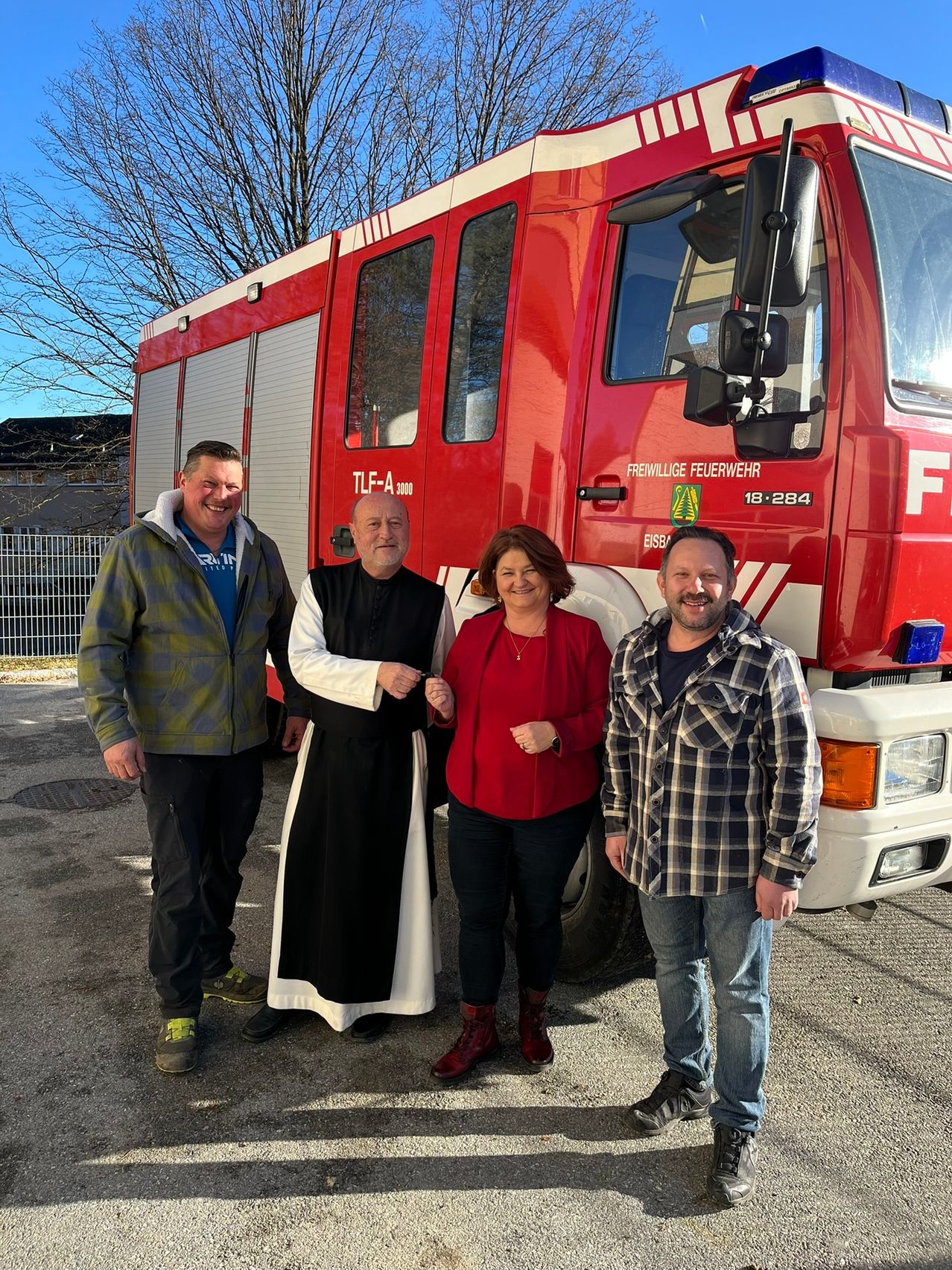 Vier Personen stehen vor einem roten Feuerwehrauto. Ein Mann und eine Frau tragen Freizeitkleidung, während ein Priester in einem schwarzen Gewand und ein weiterer Mann in einem karierten Hemd stehen.